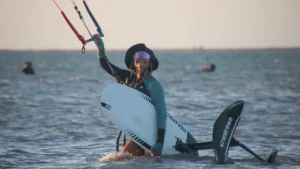 A young woman holding a hydrofoil board, with a kite flying in the background. She stands ready for an exciting kiteboarding session, demonstrating the adventurous spirit and skill of female kiteboarders.