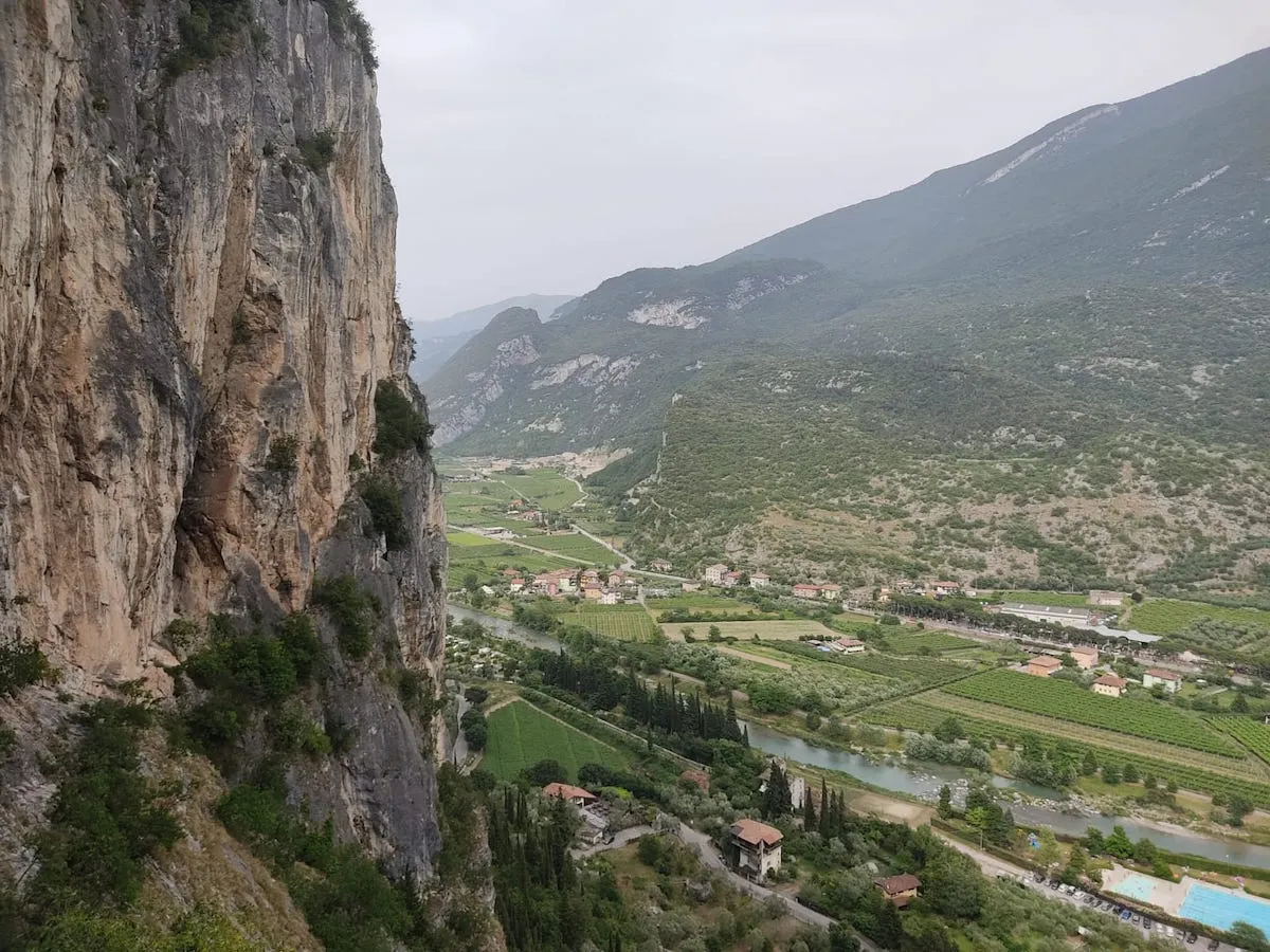 A climber scaling a rugged rock formation against the backdrop of Lake Garda, surrounded by the stunning scenery of the Italian Alps and verdant vegetation.