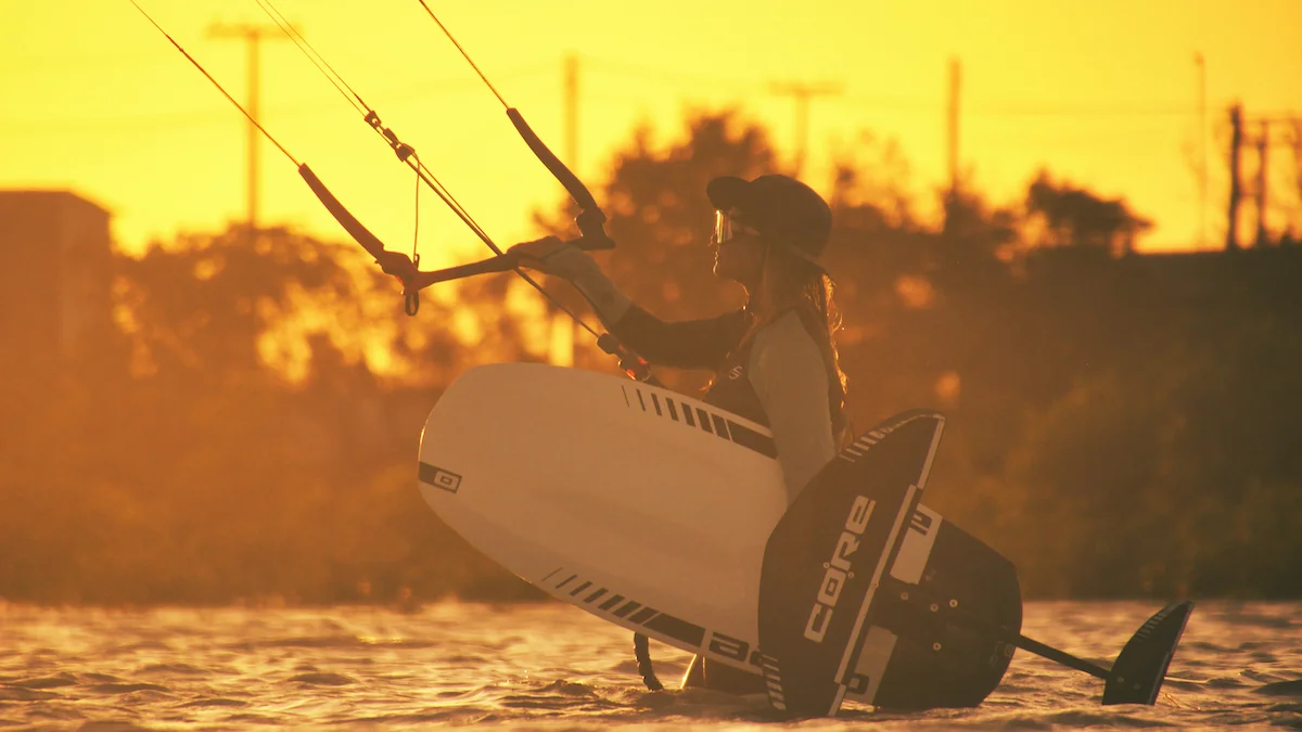 Silhouetted girl kite foiling against a vibrant sunset backdrop in Brazil, showcasing the thrill of kitecamp adventures and the beauty of the moment as she rides the waves.