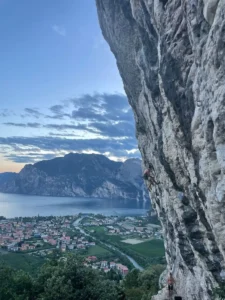 A climber ascending a rocky face near Lago di Garda, with the sparkling blue lake visible in the background, framed by towering cliffs and lush greenery.