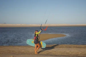 A woman walks along the shoreline, carrying her kite towards the lagoon where she will embark on a kiteboarding adventure. The anticipation of the upcoming session is palpable as she prepares to harness the wind and glide across the water with grace and skill.