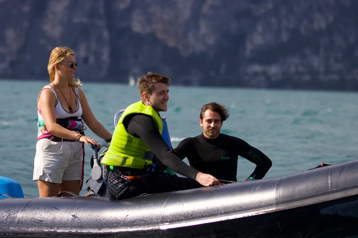 A kiteboarding instructor stands on a boat deck, surrounded by eager students, all geared up and ready to learn. The instructor gestures towards the water, providing guidance and support as they prepare for an exciting kiteboarding session on lake garda with kitevoyage
